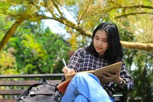 schön asiatisch jung Frau Hochschule Schüler konzentriert Schreiben auf Notizbuch und lesen Buch im draußen Stadt Park foto