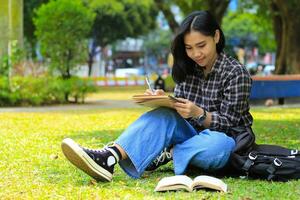 lächelnd asiatisch schön jung Frau genießen Schreiben zu tun aufführen und Idee im Notizbuch im draußen Stadt Park foto