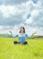 jung Frau tun Yoga auf das Gras im das Feld mit Blau Himmel im das Hintergrund, Mädchen Sitzung tun Meditation Yoga im das Feld, Frau tun Yoga draußen foto