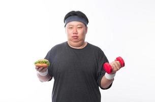 junger asiatischer lustiger fetter Sportmann, der Hamburger und Hantel hält foto