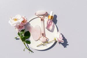 ein transparent Flasche mit ein kosmetisch Produkt mit Serum oder Rose Öl Lügen im ein Teller mit Rose Blütenblätter, ein Quarz Massagegerät, ein gua sha Schaber. oben Sicht. foto