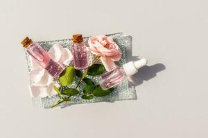ein schön Komposition von kosmetisch Flaschen mit ein Gesichts- Haut Pflege Produkt gemacht von Rose Blütenblätter. Rosa Blumen und Blütenblätter. Glas Platte. grau Hintergrund foto