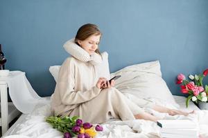 Frau sitzt im Schlafanzug auf dem Bett und tippt eine Nachricht am Telefon foto