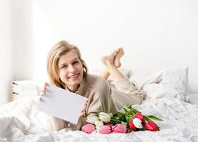 Frau liegt auf dem Bett und hält Tulpenblumenstrauß und leere Karte foto