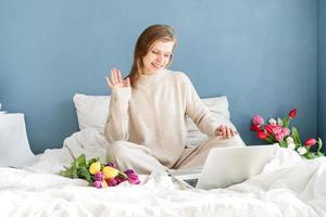 Frau sitzt im Schlafanzug auf dem Bett und plaudert am Laptop foto