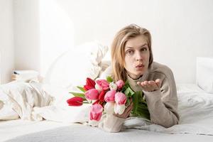 Frau liegt im Bett, hält Tulpenblumen und bläst einen Kuss foto