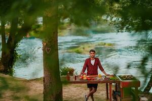 ein Mann vorbereiten ein köstlich Abendessen zum seine freunde Wer sind haben Spaß durch das Fluss im Natur foto