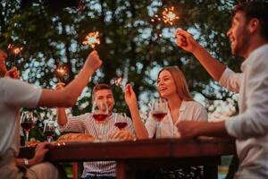 Gruppe von glücklich freunde feiern Urlaub Ferien mit Sprinkler und Trinken rot Wein während haben Picknick Französisch Abendessen Party draussen in der Nähe von das Fluss auf schön Sommer- Abend im Natur foto