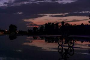 einsam Kinder Silhouette auf Fahrrad, Junge Reiten Fahrrad auf reflektierend Wasser. Hintergrund schön Sonnenuntergang. foto