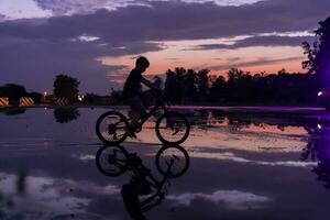 einsam Kinder Silhouette auf Fahrrad, Junge Reiten Fahrrad auf reflektierend Wasser. Hintergrund schön Sonnenuntergang. foto