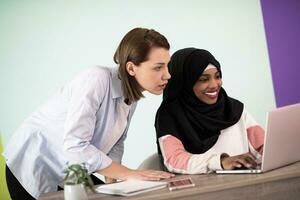 afrikanisch amerikanisch Frau mit ein Hijab und ein europäisch Frau mit ein Smartphone und Laptop im ihr Zuhause Büro foto