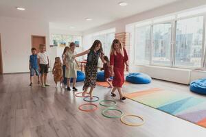 klein Kindergarten Schule Kinder mit weiblich Lehrer auf Fußboden drinnen im Klassenzimmer, tun Übung. Springen Über Hula Band Kreise Spur auf das Boden. foto