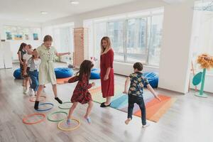 klein Kindergarten Schule Kinder mit weiblich Lehrer auf Fußboden drinnen im Klassenzimmer, tun Übung. Springen Über Hula Band Kreise Spur auf das Boden. foto