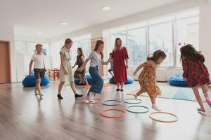 klein Kindergarten Schule Kinder mit weiblich Lehrer auf Fußboden drinnen im Klassenzimmer, tun Übung. Springen Über Hula Band Kreise Spur auf das Boden. foto