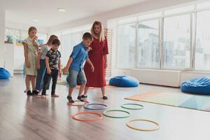 klein Kindergarten Schule Kinder mit weiblich Lehrer auf Fußboden drinnen im Klassenzimmer, tun Übung. Springen Über Hula Band Kreise Spur auf das Boden. foto