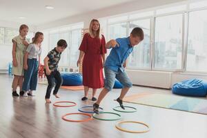 klein Kindergarten Schule Kinder mit weiblich Lehrer auf Fußboden drinnen im Klassenzimmer, tun Übung. Springen Über Hula Band Kreise Spur auf das Boden. foto