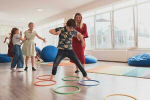 klein Kindergarten Schule Kinder mit weiblich Lehrer auf Fußboden drinnen im Klassenzimmer, tun Übung. Springen Über Hula Band Kreise Spur auf das Boden. foto