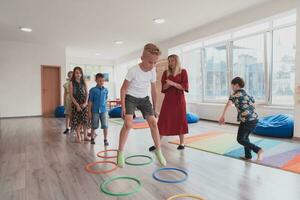 klein Kindergarten Schule Kinder mit weiblich Lehrer auf Fußboden drinnen im Klassenzimmer, tun Übung. Springen Über Hula Band Kreise Spur auf das Boden. foto