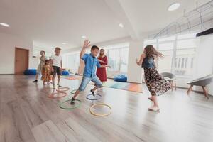 klein Kindergarten Schule Kinder mit weiblich Lehrer auf Fußboden drinnen im Klassenzimmer, tun Übung. Springen Über Hula Band Kreise Spur auf das Boden. foto