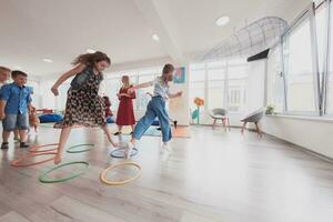 klein Kindergarten Schule Kinder mit weiblich Lehrer auf Fußboden drinnen im Klassenzimmer, tun Übung. Springen Über Hula Band Kreise Spur auf das Boden. foto