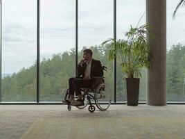 ein melancholisch Geschäftsmann im ein Rollstuhl Sitzung mit ein traurig Ausdruck, Blick durch das Fenster von ein modern Büro, vermitteln ein Sinn von Einsamkeit foto