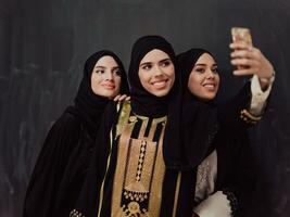 Gruppe von jung schön Muslim Frauen im modisch Kleid mit Hijab mit Smartphone während nehmen Selfie Bild im Vorderseite von schwarz Hintergrund foto
