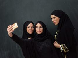 Gruppe von jung schön Muslim Frauen im modisch Kleid mit Hijab mit Smartphone während nehmen Selfie Bild im Vorderseite von schwarz Hintergrund foto