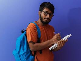 indisch Schüler mit Blau Rucksack, Brille und Notizbuch posieren auf lila Hintergrund. das Konzept von Bildung und Schulung. Zeit zu gehen zurück zu Schule foto