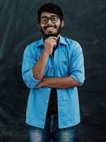 ein jung indisch Mann im ein Blau Hemd und Brille posiert nachdenklich im Vorderseite von Schule Tafel foto