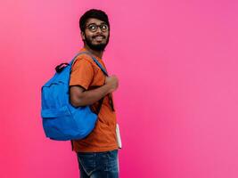 indisch Schüler mit Blau Rucksack, Brille und Notizbuch posieren auf Rosa Hintergrund. das Konzept von Bildung und Schulung. Zeit zu gehen zurück zu Schule foto