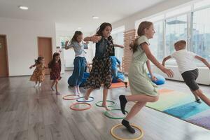 klein Kindergarten Schule Kinder mit weiblich Lehrer auf Fußboden drinnen im Klassenzimmer, tun Übung. Springen Über Hula Band Kreise Spur auf das Boden. foto