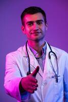 Porträt von ein Held im ein Weiß Mantel. heiter lächelnd jung Arzt mit Stethoskop im medizinisch Krankenhaus Stehen auf Blau und Rosa Hintergrund. Warnung Über das Achtung von das Coronavirus covid-19 foto