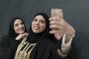 Gruppe von jung schön Muslim Frauen im modisch Kleid mit Hijab mit Smartphone während nehmen Selfie Bild im Vorderseite von schwarz Hintergrund foto