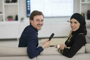 jung Muslim Paar Frau tragen islamisch Hijab Kleider Sitzung auf Sofa Aufpassen Fernseher zusammen während das Monat von Ramadan beim modern Zuhause foto