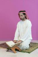 jung arabisch Muslim Mann im traditionell Kleider lesen heilig Buch Koran auf das beten Teppich Vor iftar Abendessen während ein Ramadan Fest beim Zuhause foto