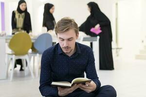 jung Muslim Mann Sitzung auf das Fußboden während lesen heilig Buch Koran Vor iftar Abendessen während ein Ramadan Fest beim Zuhause foto