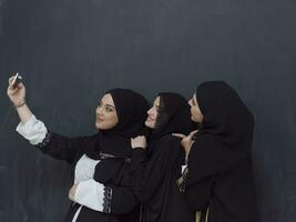 Gruppe von jung Muslim Frauen im modisch Kleid mit Hijab mit Smartphone während nehmen Selfie Bild im Vorderseite von schwarz Hintergrund foto