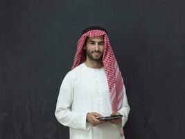 jung arabisch Muslim Geschäftsmann mit Tablette Computer tragen Hijab Kleider im Vorderseite von schwarz Tafel foto