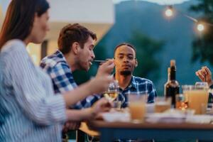 ein Gruppe von jung vielfältig Menschen haben Abendessen auf das Terrasse von ein modern Haus im das Abend. Spaß zum freunde und Familie. Feier von Feiertage, Hochzeiten mit Grill. foto