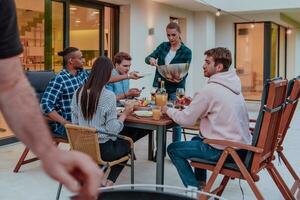 ein Gruppe von jung vielfältig Menschen haben Abendessen auf das Terrasse von ein modern Haus im das Abend. Spaß zum freunde und Familie. Feier von Feiertage, Hochzeiten mit Grill. foto