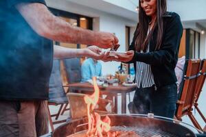 ein Gruppe von freunde und Familie Grill zusammen im das Abend auf das Terrasse im Vorderseite von ein groß modern Haus foto