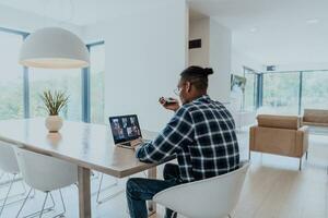 afrikanisch amerikanisch Mann im Brille Sitzung beim ein Tabelle im ein modern Leben Zimmer, mit ein Laptop und Smartphone zum Geschäft Video Plaudern, Konversation mit freunde und Unterhaltung foto