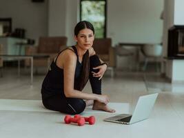 ein jung Frau im Sportbekleidung ist Sitzung im das Leben Zimmer und vorbereiten zum online Ausbildung während mit ein Laptop foto