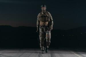 Heer Soldat im Kampf Uniformen mit ein Angriff Gewehr, Teller Träger und Kampf Helm gehen auf ein gefährlich Mission auf ein regnerisch Nacht. foto