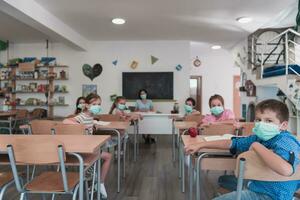 gemischtrassig Gruppe von Kinder tragen Gesicht Masken Arbeiten beim Klasse, Schreiben und Hören Erklärungen von Lehrer im Klassenzimmer foto