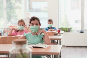 gemischtrassig Gruppe von Kinder tragen Gesicht Masken Arbeiten beim Klasse, Schreiben und Hören Erklärungen von Lehrer im Klassenzimmer foto