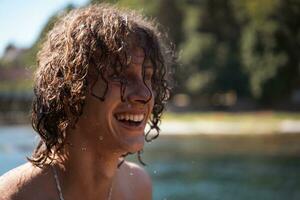 Porträt von ein jung Teen Junge mit lockig nass Haar in der Nähe von das Fluss haben Spaß mit freunde beim ein Sommer- Party. foto