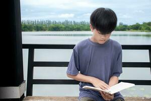 asiatisch jung Junge lesen Buch auf Stuhl beim Flussufer Hintergrund ist Wald und Himmel mit Sonnenaufgang, Junge Blau Hemd, Kinder mit Buch, Konzept Lernen und Bildung, natürlich schön Hintergrund. foto