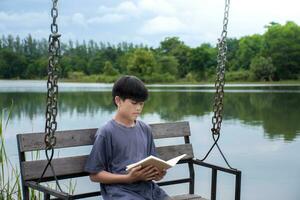 asiatisch Junge Sitzung lesen Buch auf schwingen beim Flussufer Hintergrund ist Wald und Himmel mit Sonnenaufgang, Junge Blau Hemd, Kinder mit Buch, Konzept Lernen und Bildung, natürlich schön Hintergrund. foto