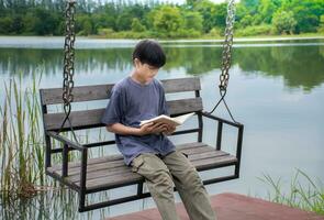 asiatisch jung Junge lesen Buch auf schwingen beim Flussufer Hintergrund ist Wald und Himmel mit Sonnenaufgang, Junge Blau Hemd, Kinder mit Buch, Konzept Lernen und Bildung, natürlich schön Hintergrund. foto